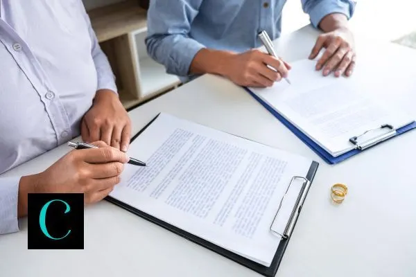 Two people sitting at a desk signing divorce papers with two wedding rings placed nearby.