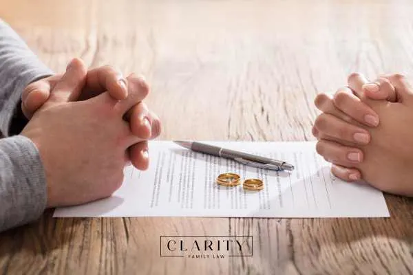 A pen and two wedding rings on a divorce agreement document, with the hands of a couple on either side of the table.