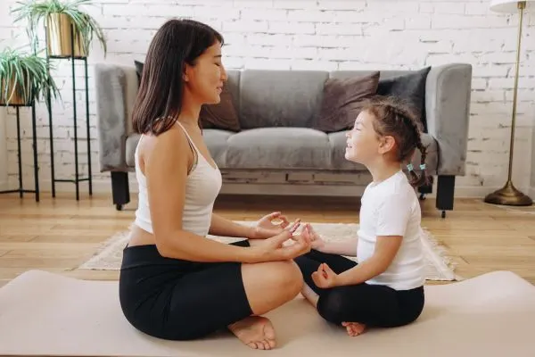 A woman and a young girl sitting cross-legged on a yoga mat.