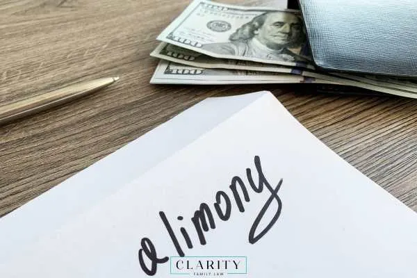 envelope with the word "alimony" written on it, sits on a desk next to a pen and a walletwith US dollar bills.
