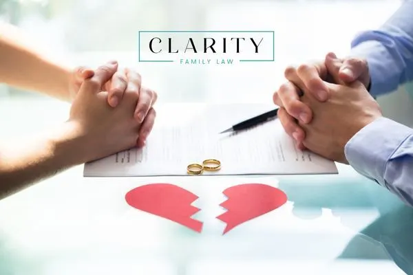 hands clasped over a document featuring two wedding rings and a pen, with a paper cutout of a broken heart