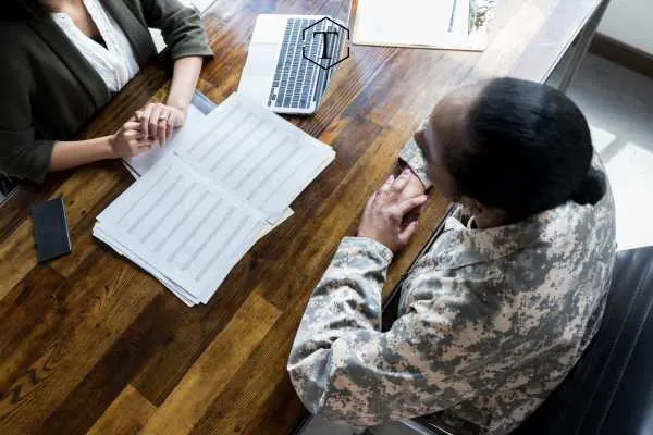 Service member and civilian consult over documents and a laptop.