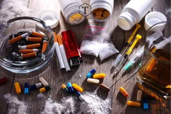 An overhead shot of an assortment of illicit drugs, drug paraphernalia, and alcoholic beverages on a wooden table.