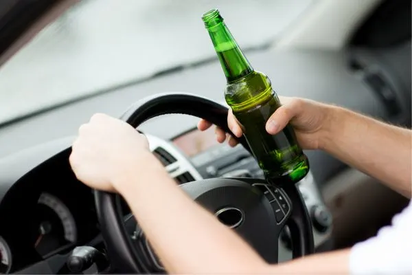 Hands holding a beer bottle while driving a car, implying drunk driving.