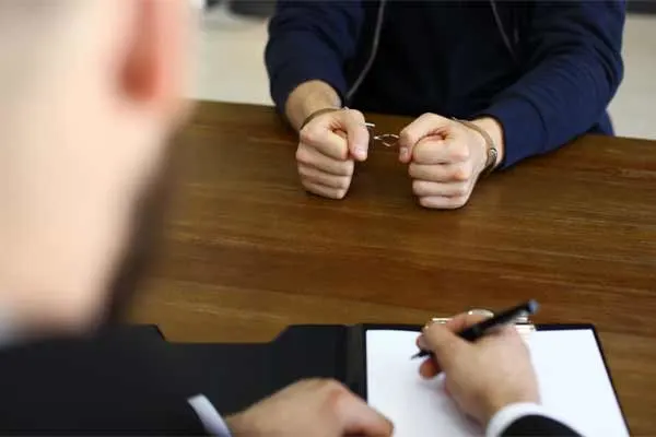A close-up shot of a person in handcuffs sitting across a table from another person who is taking notes on a clipboard.