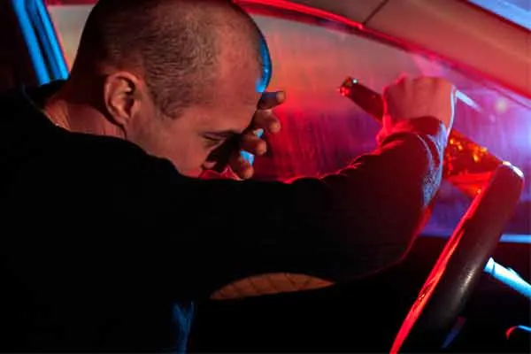 man sits slumped over the steering wheel of a car, with one hand on his forehead, illuminated by flashing red and blue lights.