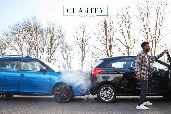 A man opens the door of a black SUV that has rear-ended a blue car, which is emitting smoke from its front. The accident scene is on a road with bare trees in the background. The "Clarity Family Law" logo is at the top.