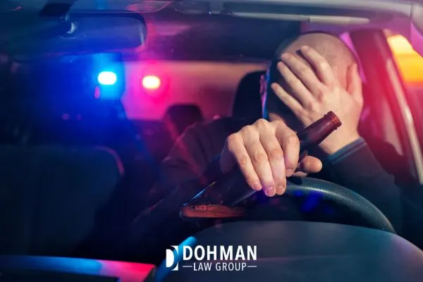 Man covering face in car with police lights, holding a bottle.