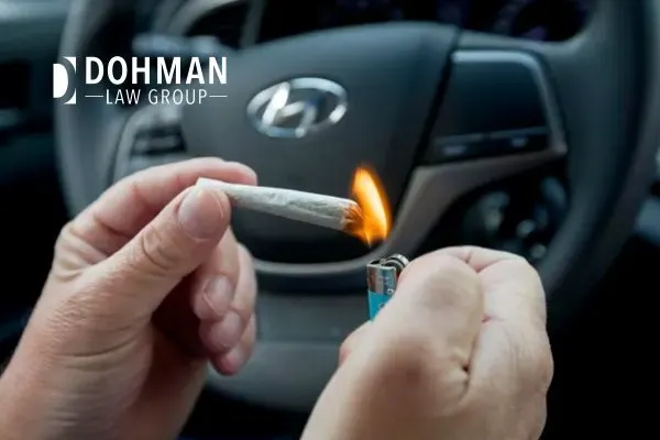 A person's hands are lighting a marijuana joint with a lighter, with a car's steering wheel in the background.
