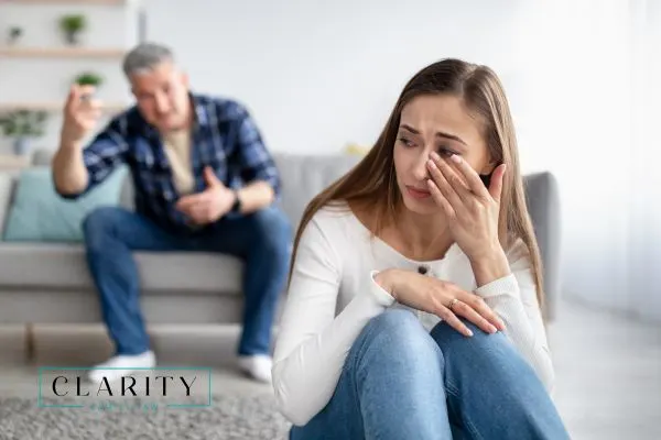 A woman sits on the floor, crying and wiping her eye, while a man in the background gestures angrily.