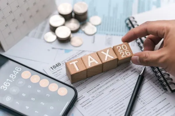 A hand placing wooden blocks that spell "TAX 2026" on top of a 1040 tax form surrounded by coins and a calculator.