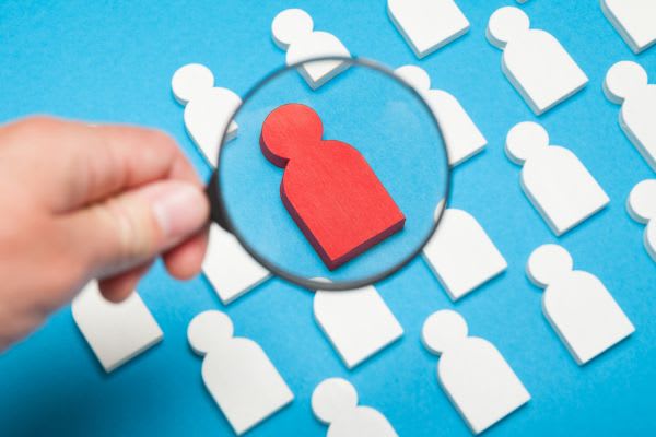 A magnifying glass over a red person icon in a crowd of white person icons, on a blue background.