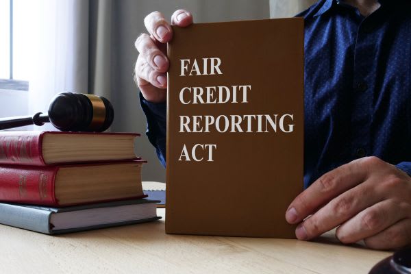 A person holding up a book titled "FAIR CREDIT REPORTING ACT," with legal books and a gavel on a desk beside it.