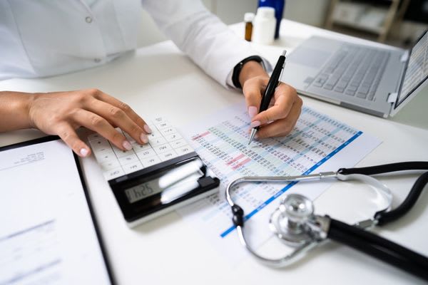 A person uses a calculator and pen to review financial documents next to a stethoscope