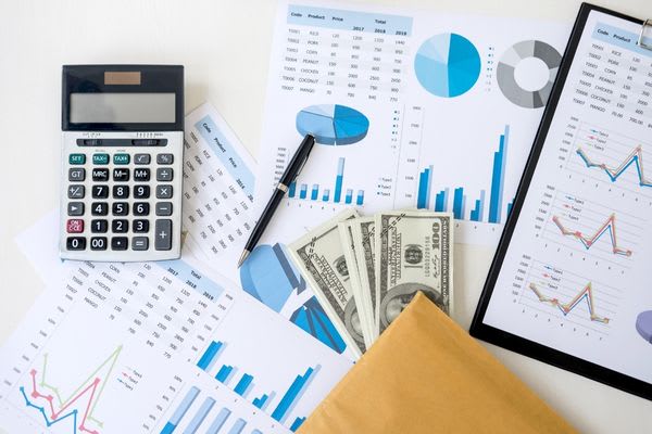 An overhead view of financial spreadsheets, surrounded by a calculator, a pen, and a stack of $100 bills