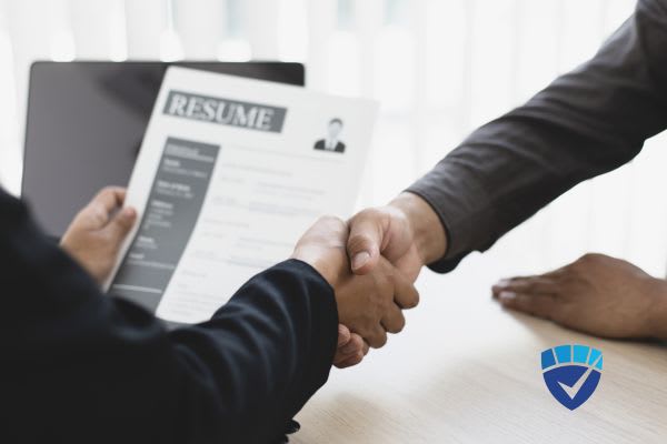 Two people shaking hands over a desk while one holds a professional resume.