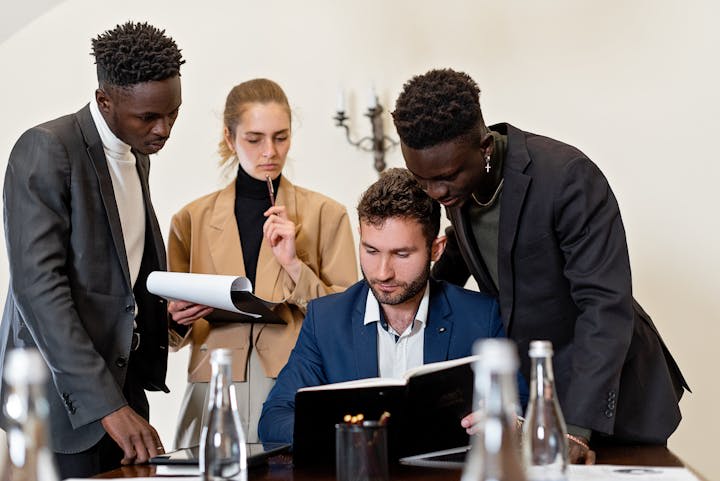 professionals in business attire gather around a table, leaning in to review a notebook