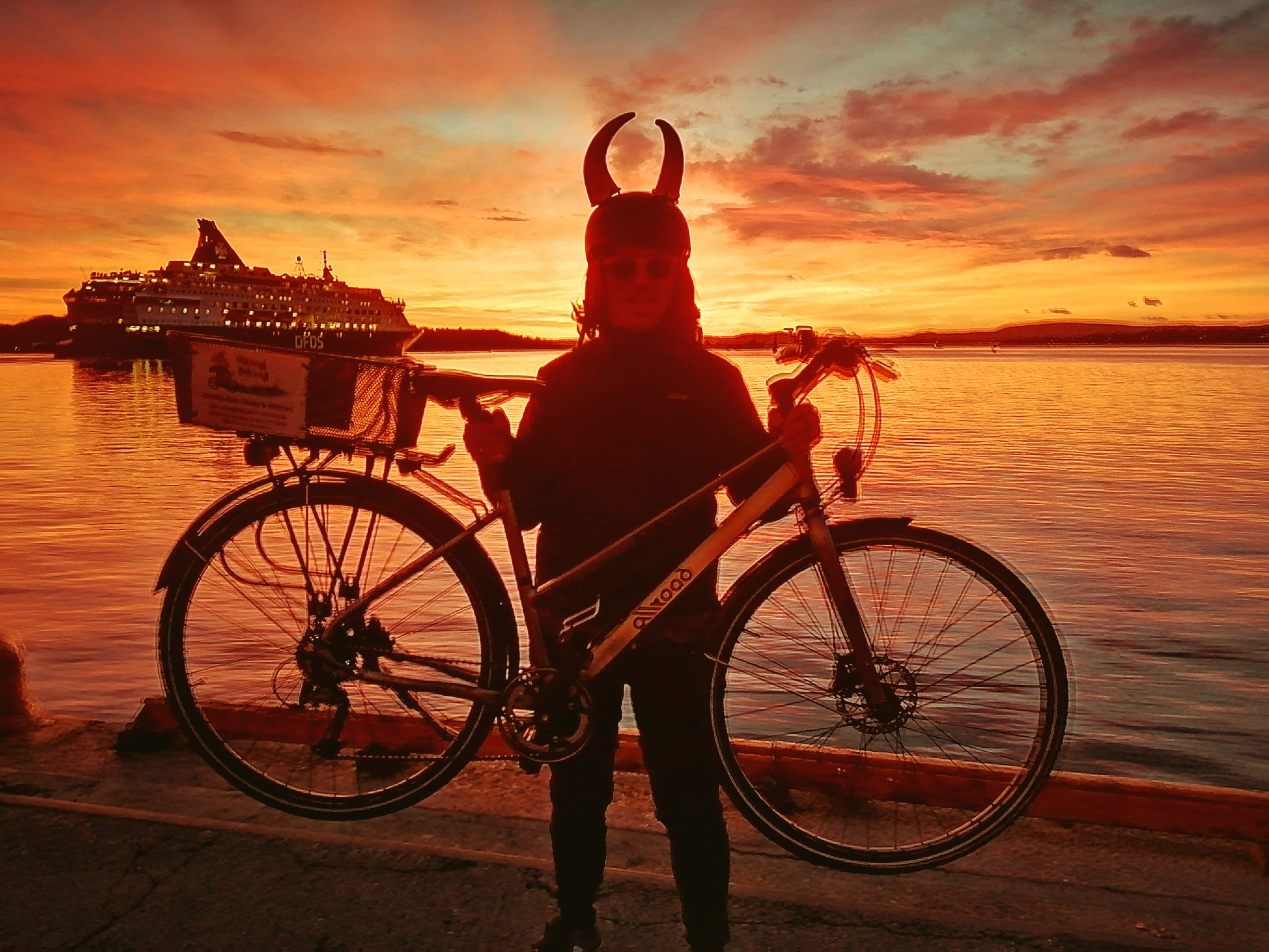A man wearing a viking helmet while lifting his bike. A vivid red sunset is playing out in the background.