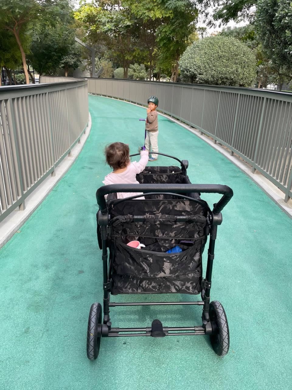 Older child riding a scooter while a younger sibling rides comfortably in a STROLLZ. stroller wagon.