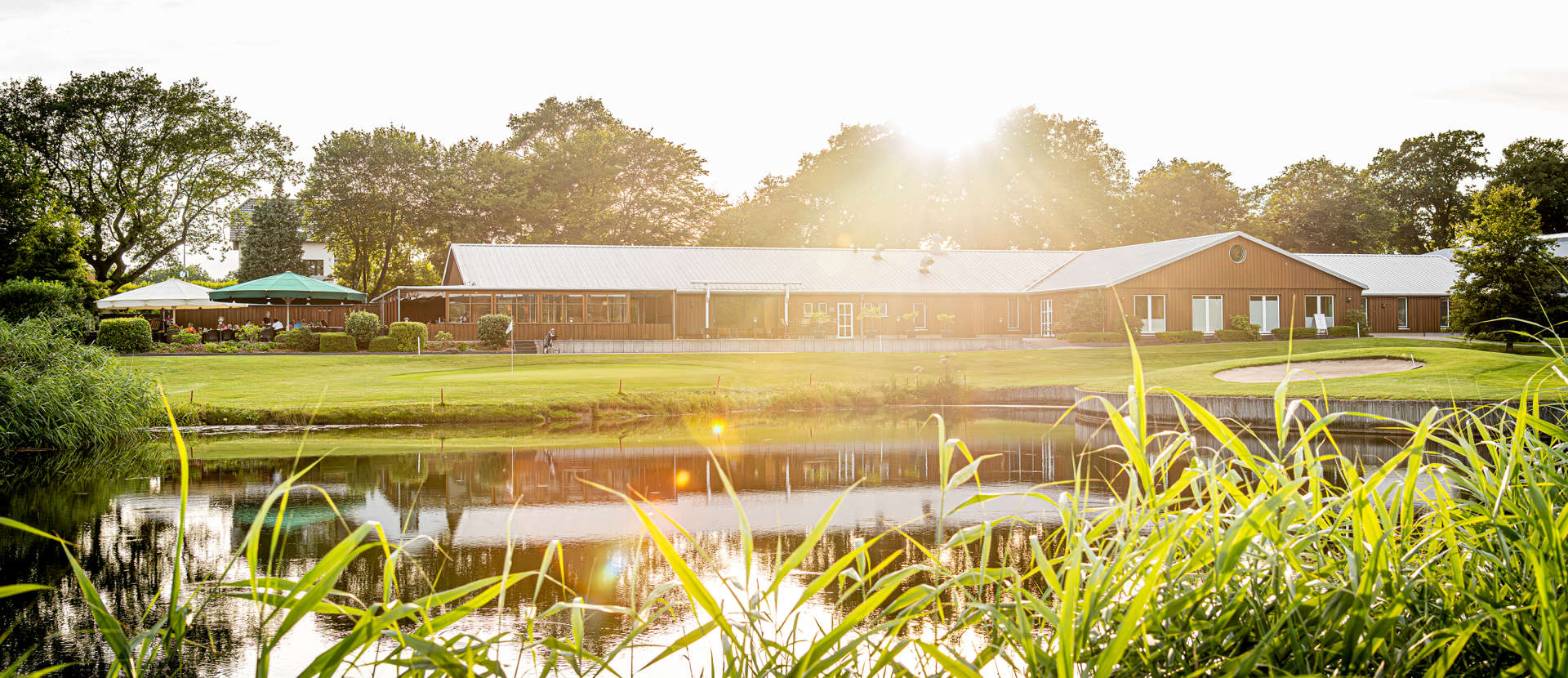Ganzheitliche Ansicht unseres Clubhauses im Golf Gut Glinde mit einladender Sonnenterrasse und dem malerischen Teich der Bahn 9 direkt vor dem Gebäude. Das stilvolle Clubhaus bietet eine perfekte Kombination aus modernem Komfort und Naturidylle, ideal für entspannte Pausen, gesellige Runden oder gepflegte Dinner nach dem Golfspiel. Der Teich und die grüne Umgebung schaffen eine ruhige Atmosphäre und bieten den perfekten Blick auf die gepflegten Fairways unseres 18-Loch-Golfplatzes.