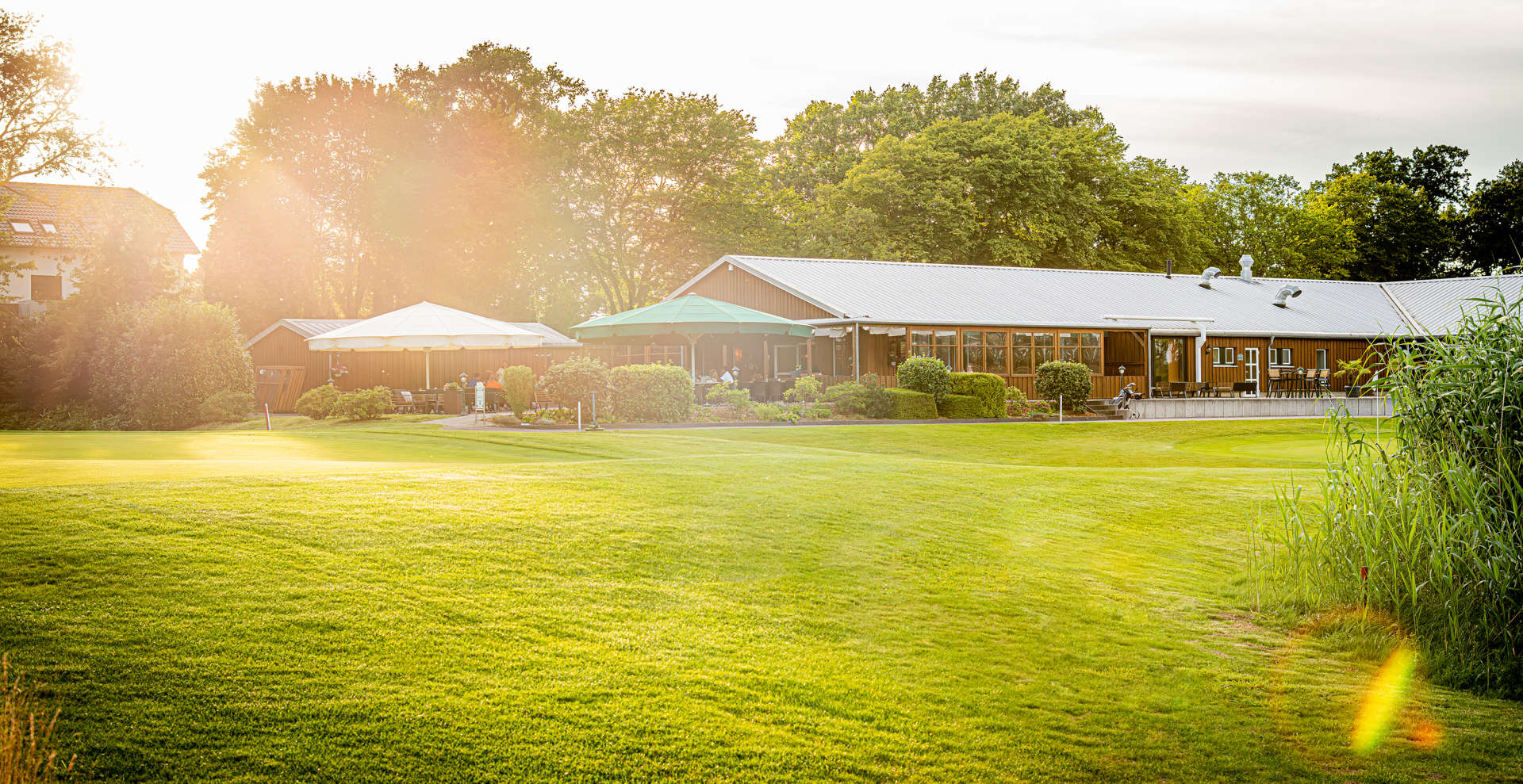 Ansicht unseres Clubhauses im Golf Gut Glinde, inklusive einladender Sonnenterrasse. Das moderne Clubhaus bietet den perfekten Rahmen für Mitgliedertreffen, entspannte Auszeiten nach dem Golfen und kulinarische Genüsse in stilvollem Ambiente. Die großzügige Terrasse lädt zum Verweilen im Freien ein, mit Blick auf den gepflegten Golfplatz und die idyllische Natur – ideal für Golfer und Gäste, die eine angenehme Zeit in einer herzlichen Atmosphäre verbringen möchten.