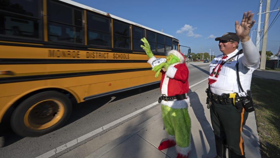 En esta foto proporcionada por Florida Keys News Bureau, el coronel de la oficina del alguacil del condado de Monroe, Lou Caputo, a la izquierda, disfrazado de Grinch, y el diputado Andrew Leird, a la derecha, saludan a un autobús escolar que circula por la autopista Florida Keys Overseas Highway el martes 13 de diciembre. de 2022, en Marathon, Florida. Cuando los conductores son detenidos por exceso de velocidad en una zona escolar, Caputo les ofrece elegir entre una cebolla o una multa de tránsito. Es una tradición navideña en los Cayos que Caputo comenzó hace 20 años para educar a los conductores que las escuelas de los Cayos permanecen abiertas hasta el 16 de diciembre. (Andy Newman/Florida Keys News Bureau vía AP)