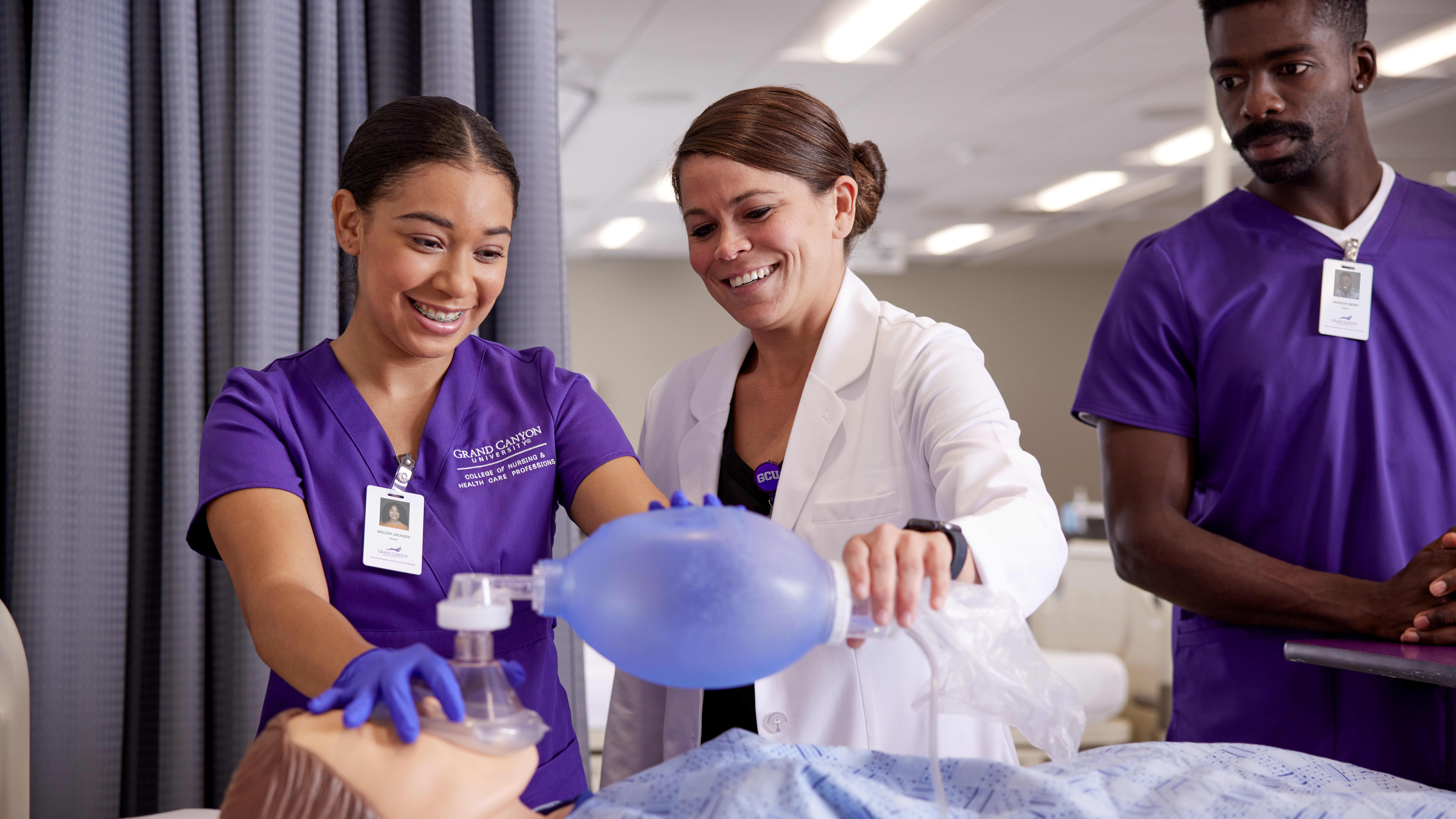 Group of Boise (Meridian, ID) ABSN students in purple scrubs surround a fake patient in a hospital bed
