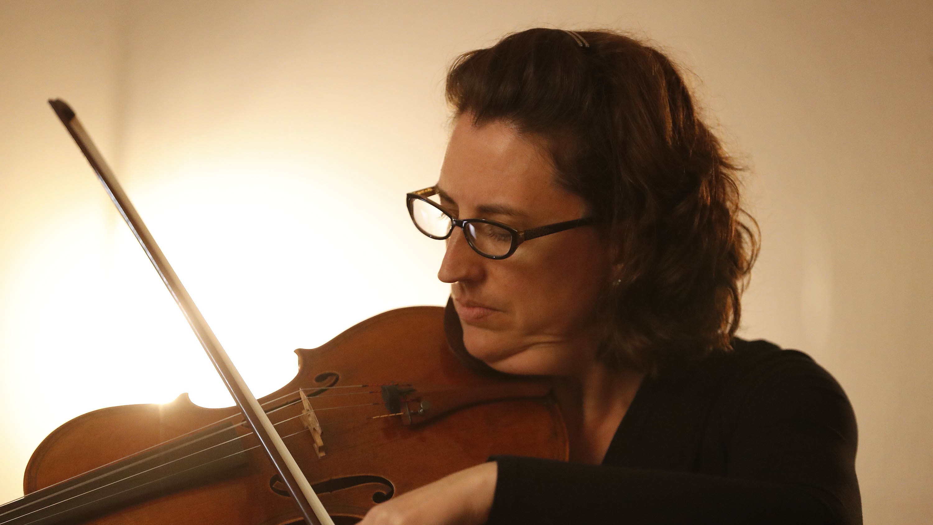Brunette female with medium length curly hair and black plastic frame glasses plays violin at faculty recital