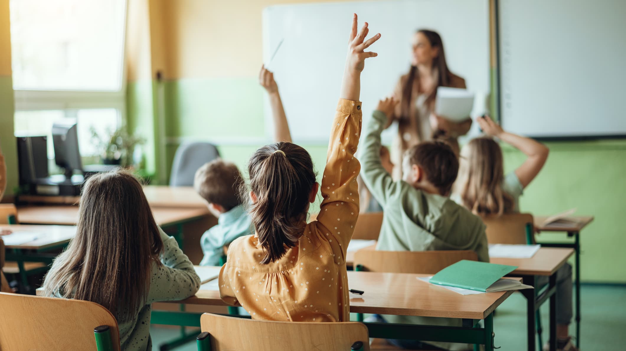 Shot of Elementary Students Backs Raising Hands at Desk in a Classroom With Teacher at the Front of Classroom Blurred and Out of Focus