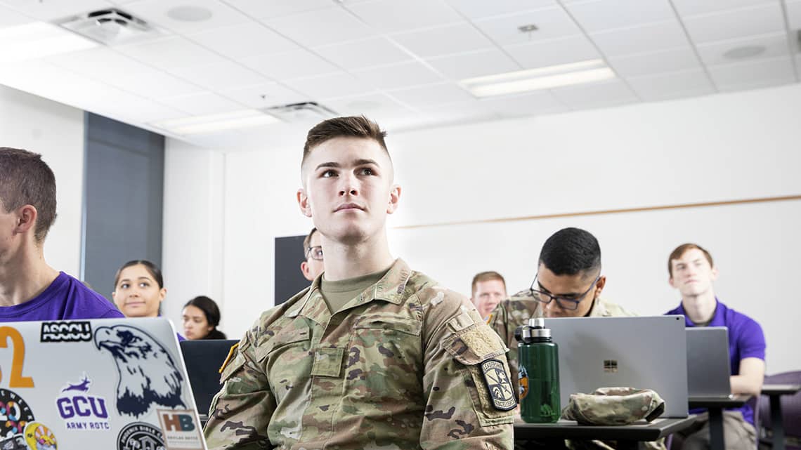 Male ROTC student in fatigues gazes over at professor in class with laptop open on desk