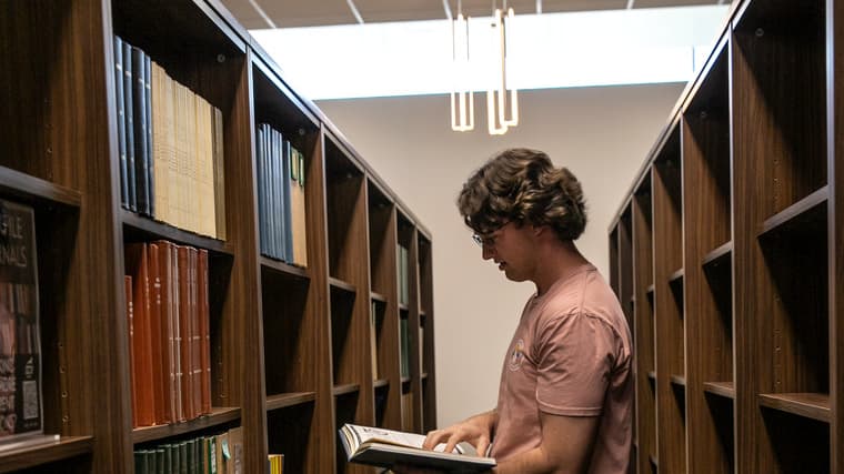 White male in a light pink shirt and darker shorts stands reading a book in GCTS
