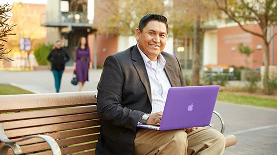 Male adult student sitting on a bench working on his laptop