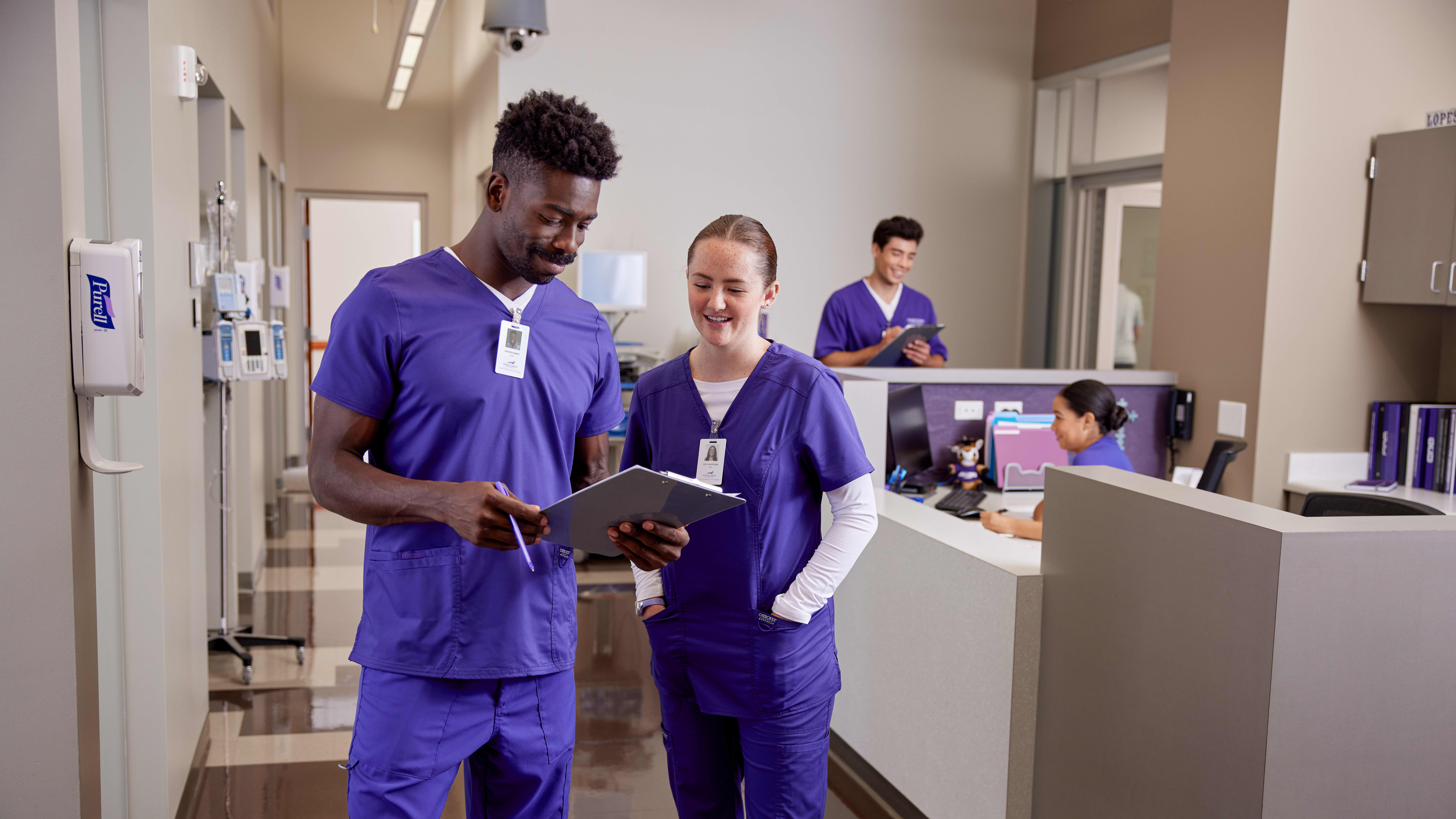 Two New Mexico ABSN students in purple scrubs walk past a nurse's station