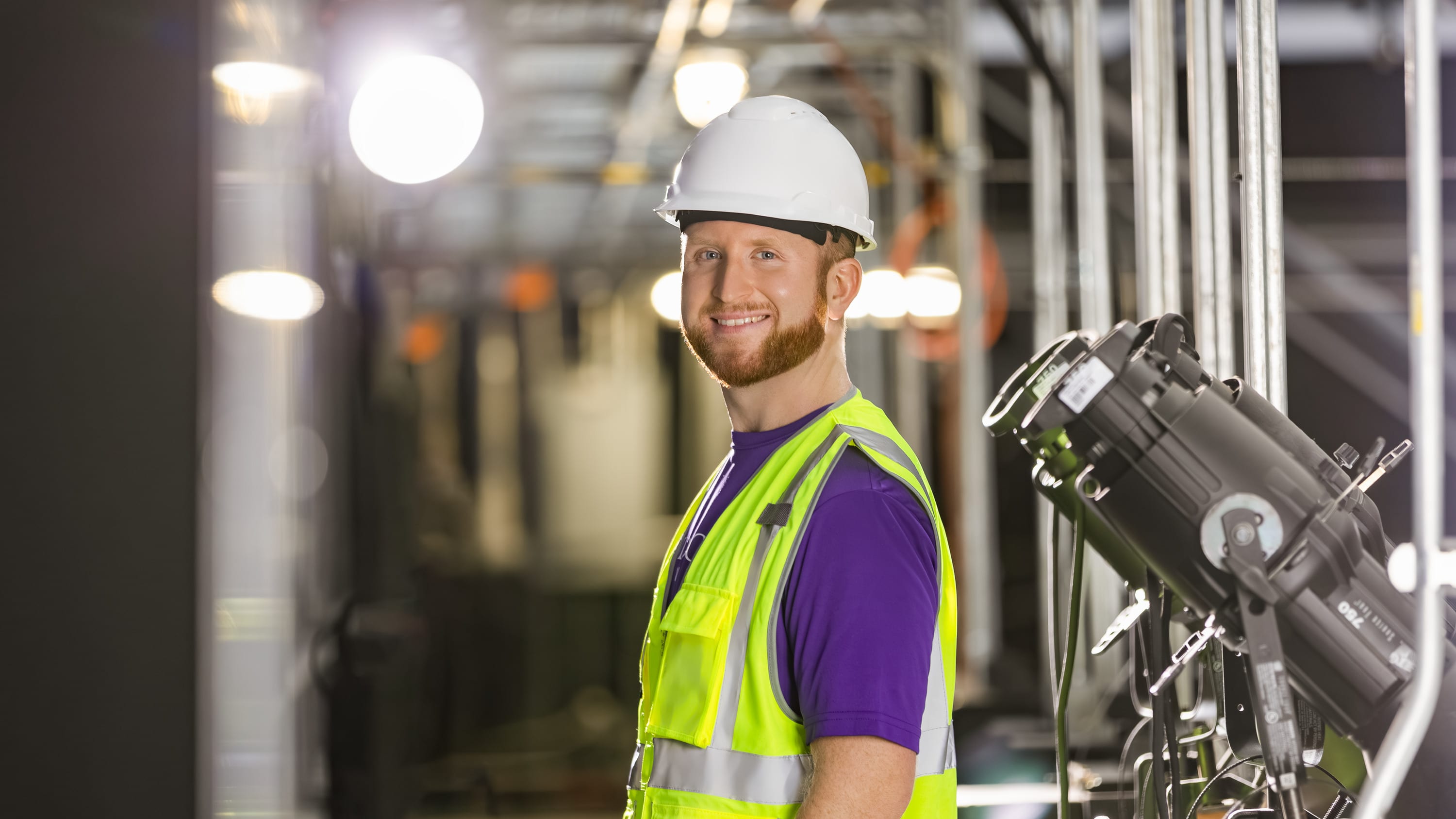 Man in industrial setting standing with purple shirt and hard hat