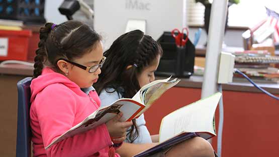 Two young elementary-aged children reading books on chairs