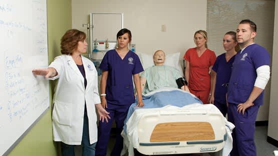 Middle-aged female nurse educator breaks down diagnosis on whiteboard while nurse students surround a dummy patient's bed