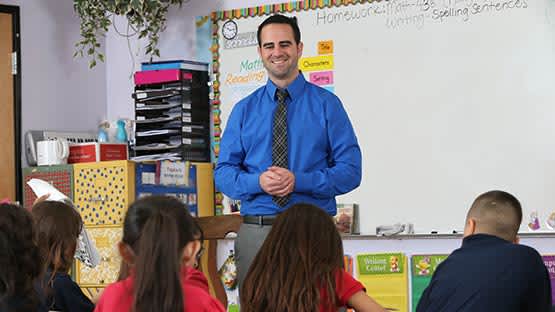 Hispanic male teacher stands in front of classroom whiteboard with blue long sleeved shirt on with third graders in school uniforms facing him