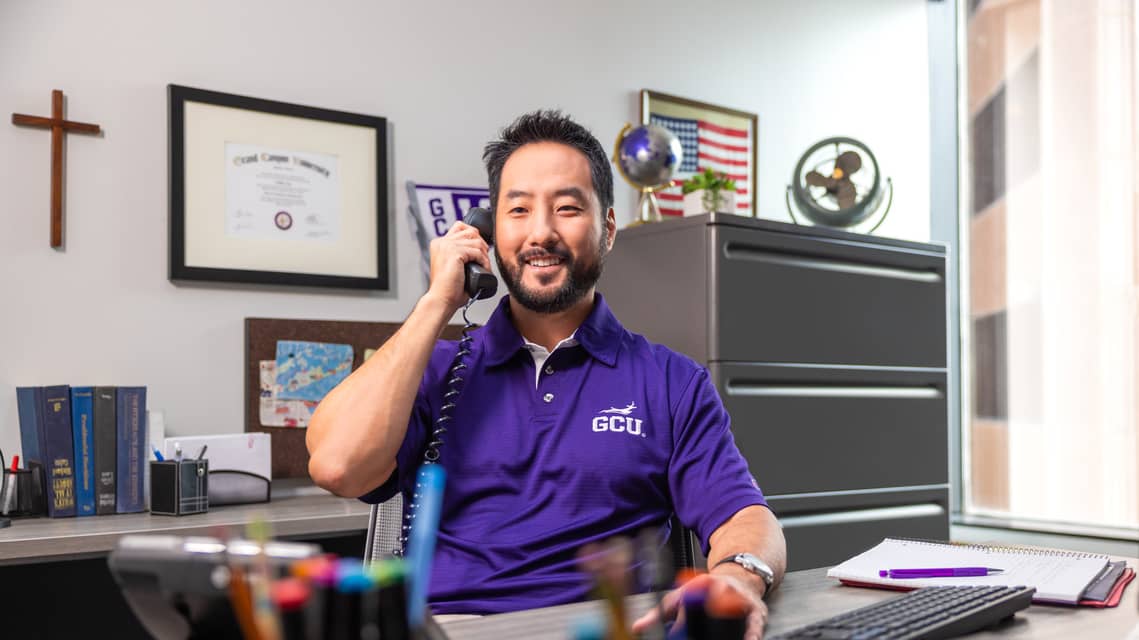 Asian male student university counselor wearing a purple GCU polo answers a student call about tuition