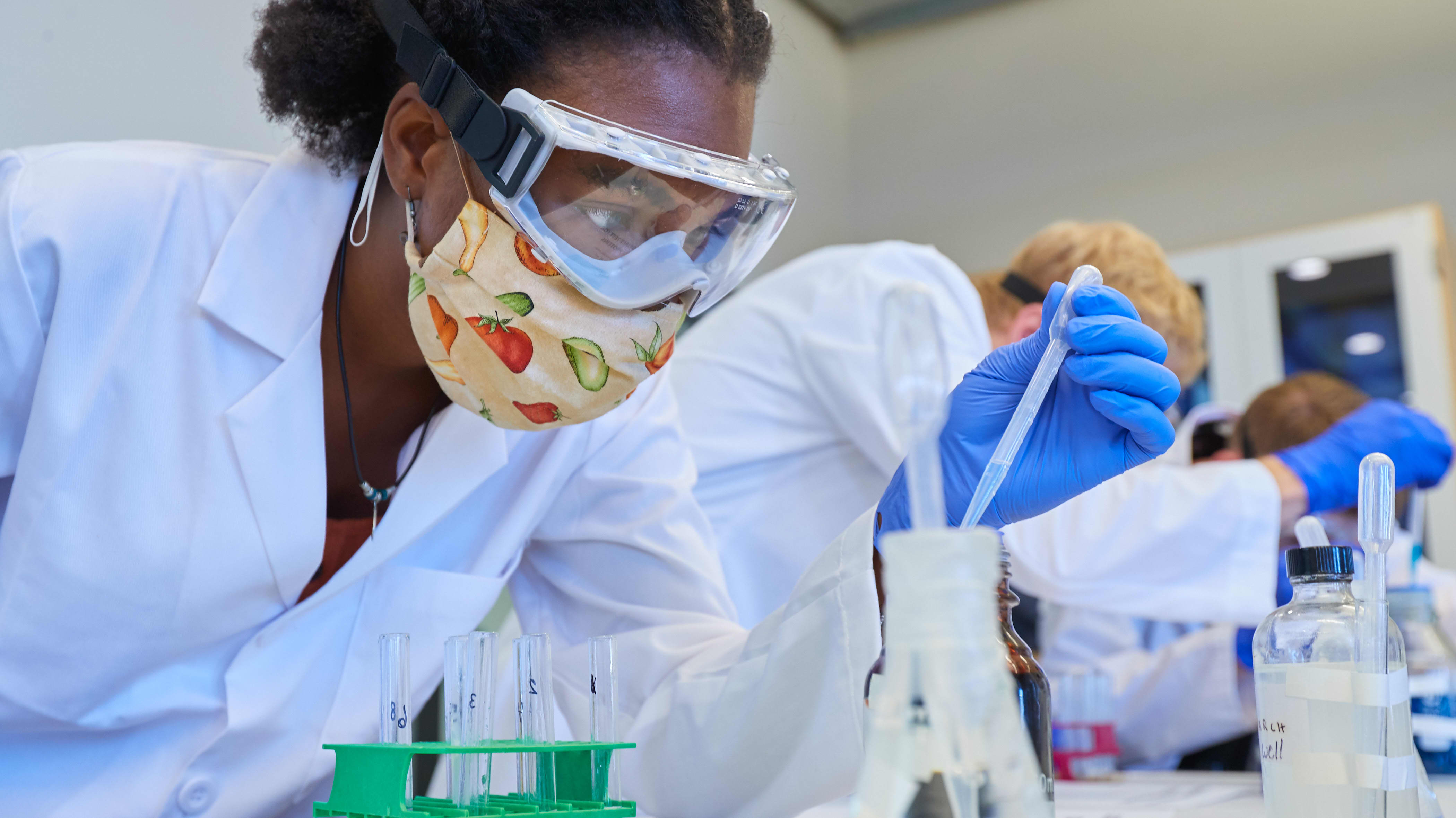 Female student wearing white lab coat and safety glasses looking at sample in the lab