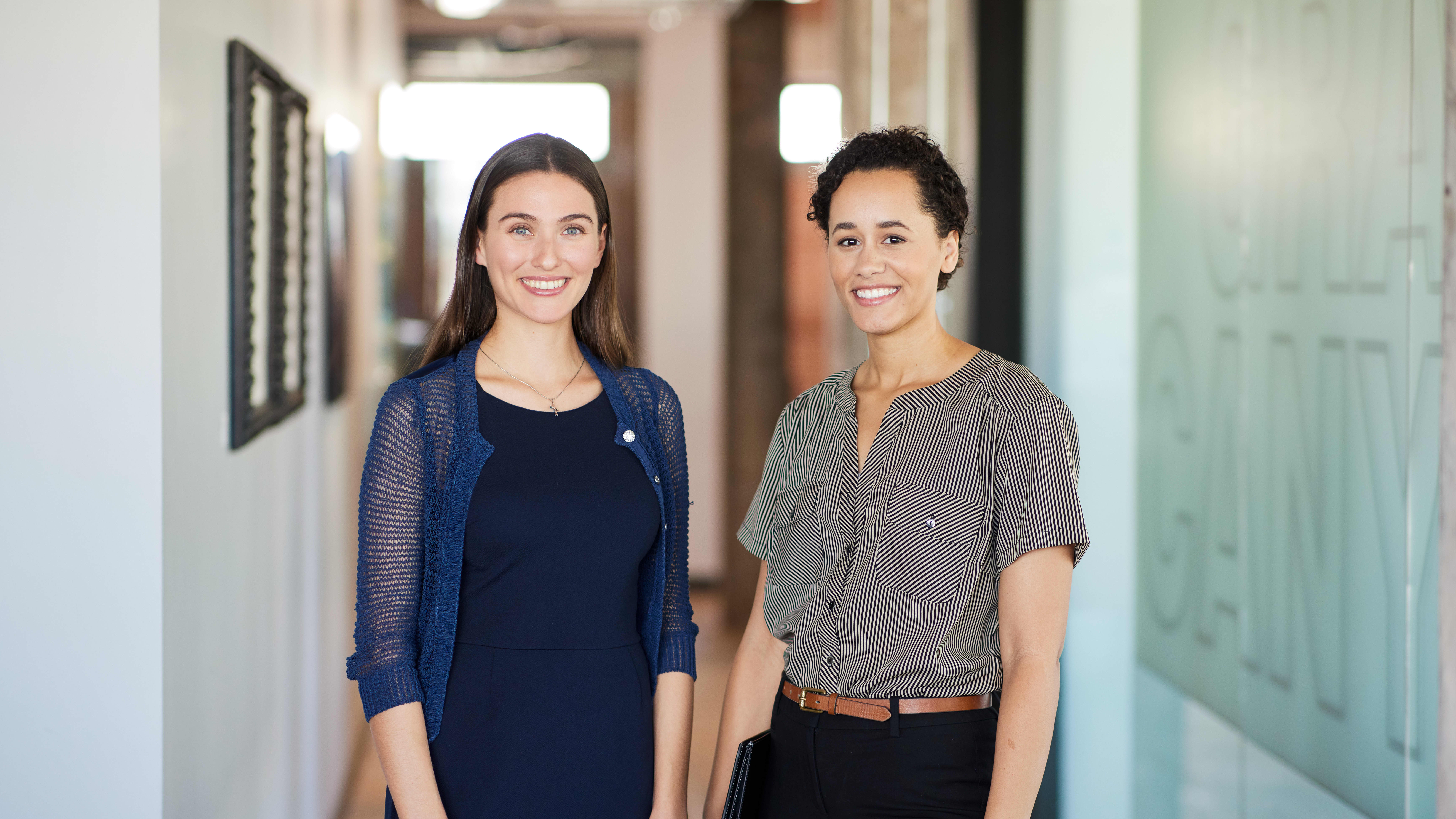 Two women in business casual attire smiling in hallway