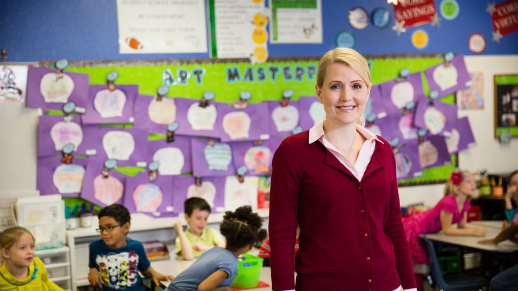 Teacher standing in front of classroom smiling at camera with students working in background