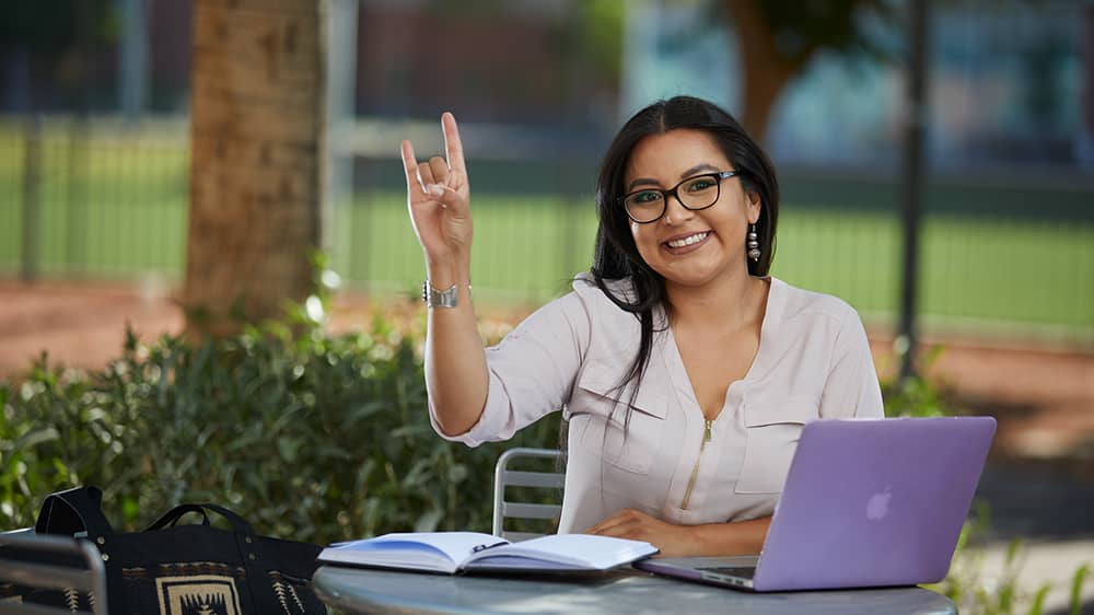 Master's student sitting with laptop holding up lopes sign