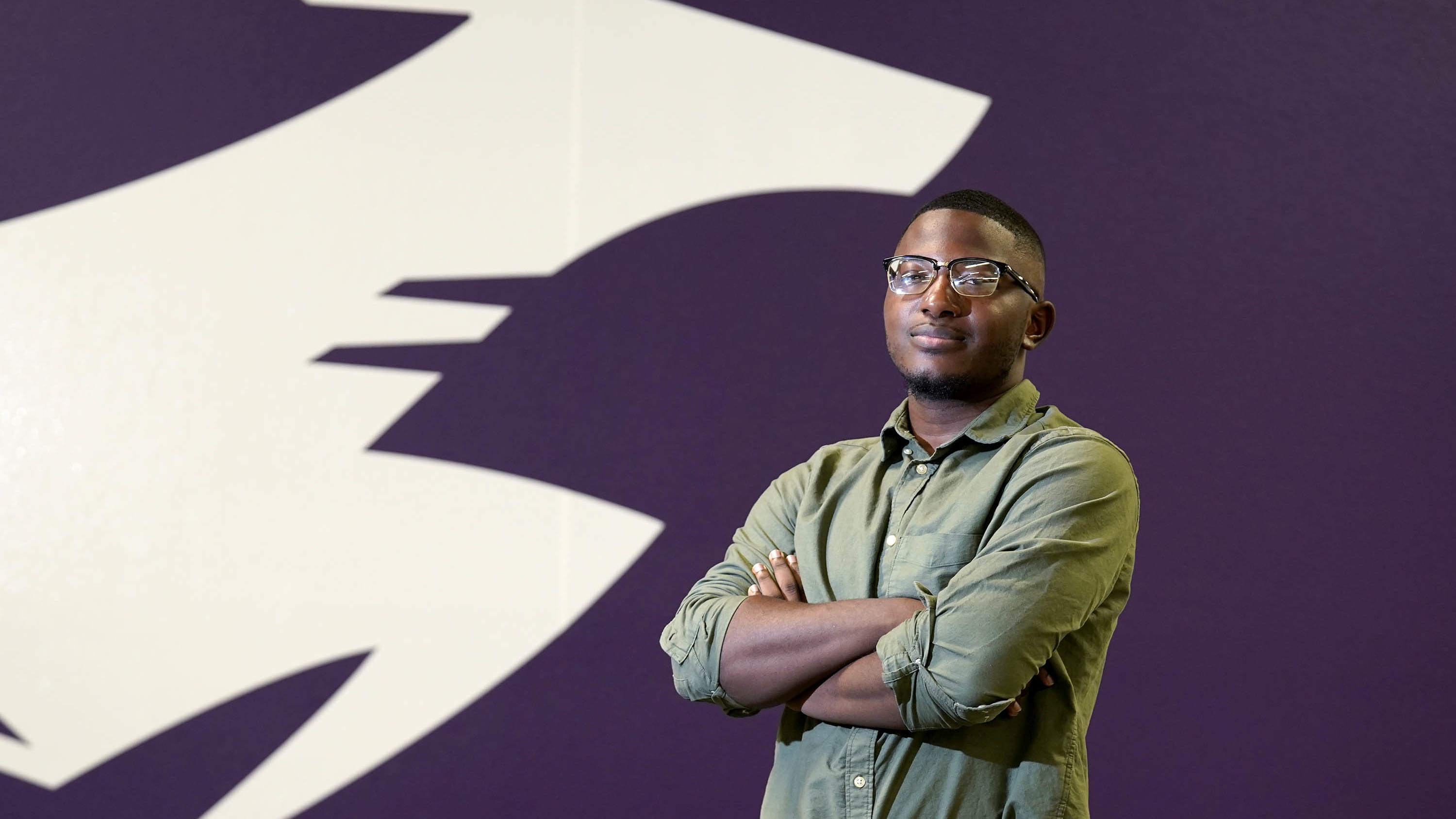 Male fine arts degree major smiling and standing in front of purple GCU wall