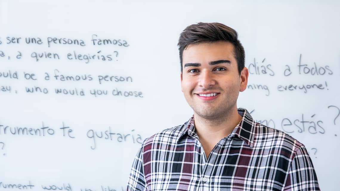 Hispanic male teacher in plaid button down stands in front of white board with Spanish language written on it