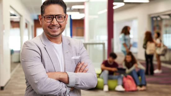 Hispanic young professor with trendy black frames stands with crossed arms in a pale gray suit with students sitting and standing in background