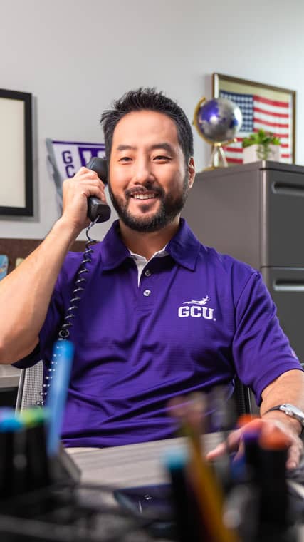Asian male student university counselor wearing a purple GCU polo answers a student call about tuition