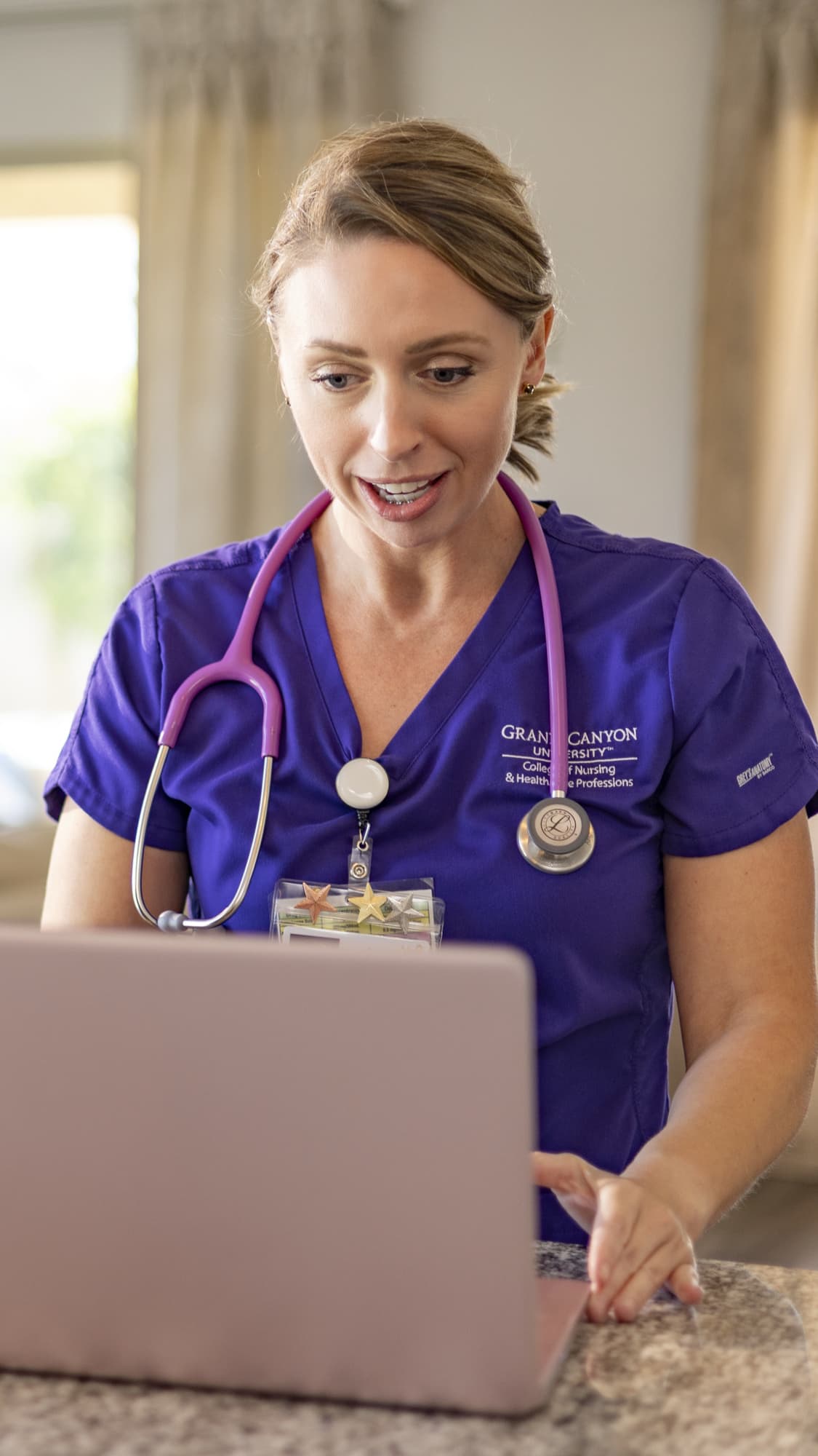 Woman in scrubs using laptop in home kitchen to earn BSN degree online
