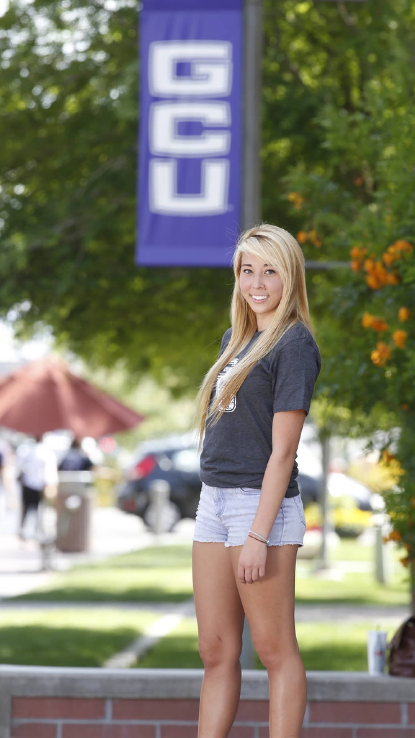 Slender blonde female GCU student stands near a retaining wall tree and a pole with a GCU feather banner