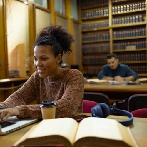 Happy student working in a library with an open book and laptop