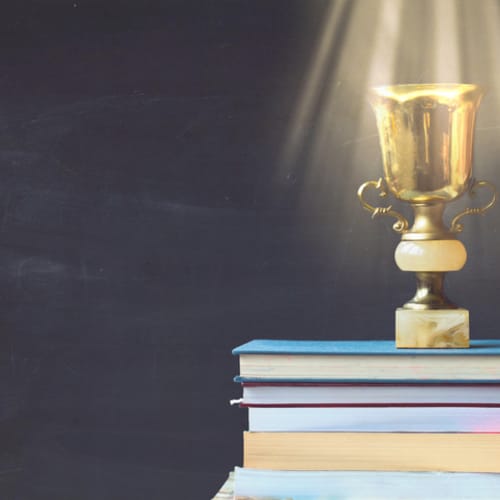 Golden trophy on pile of books, against blackboard, with sun rays over trophy; learning/achievement concept - stock photo